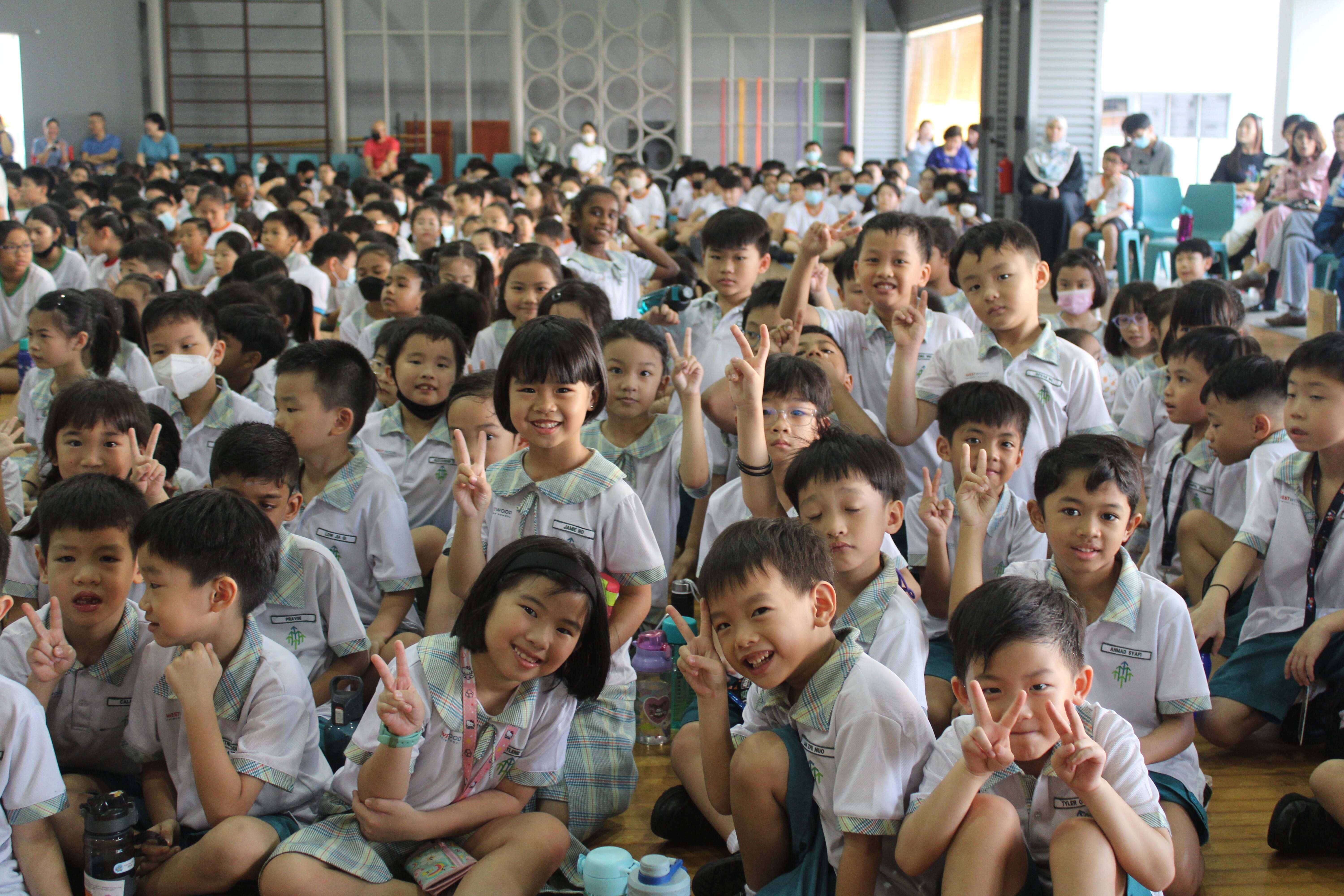 Crowd of schoolchildren in white uniforms sitting indoors, many smiling and holding up peace signs.